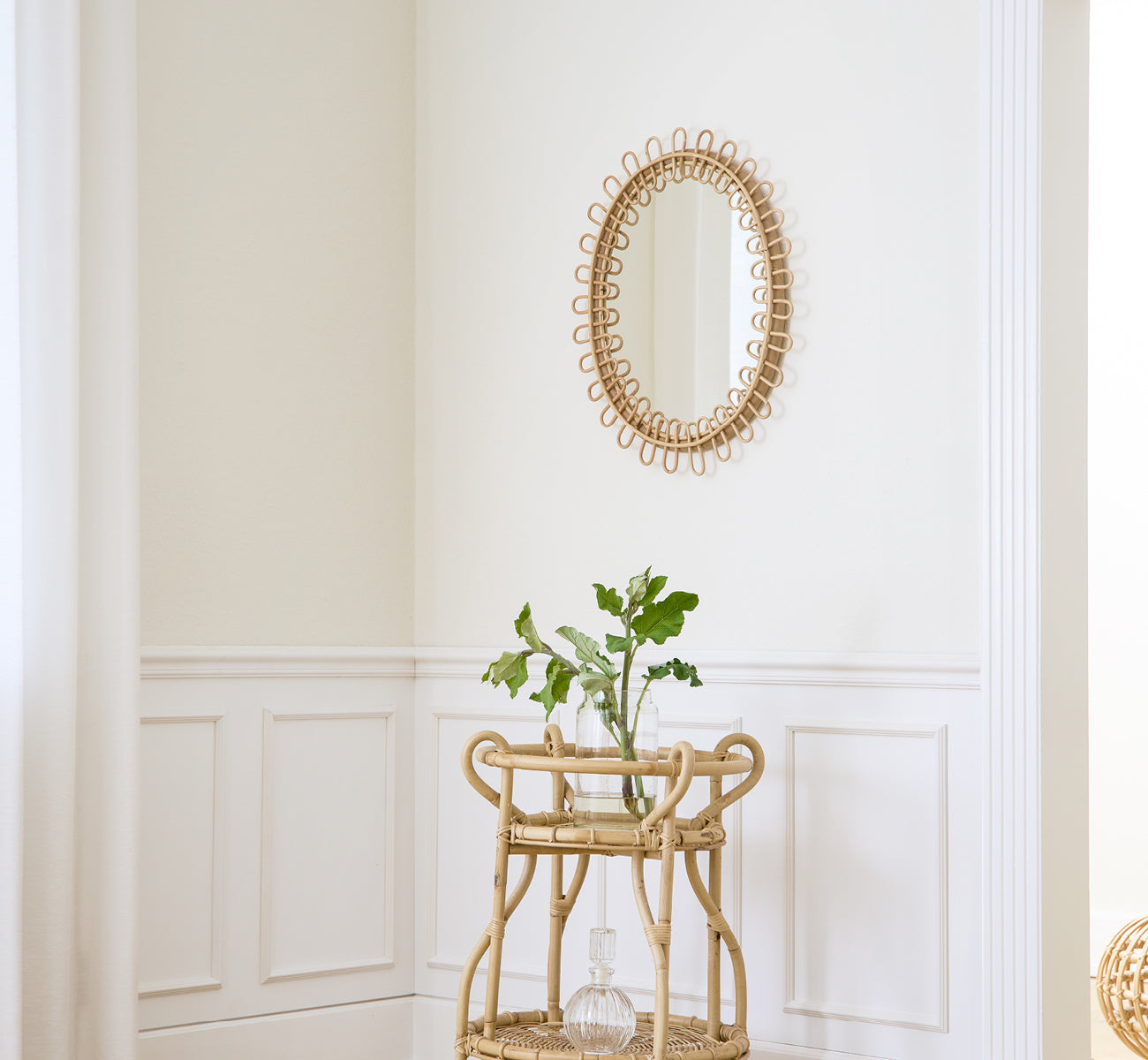 Dies ist ein Lifestyle-Bild von Luella Spiegel in polished natural rattan. An einer weiß vertäfelten Wand über einem Rattan-Barwagen mit Grün montiert.
