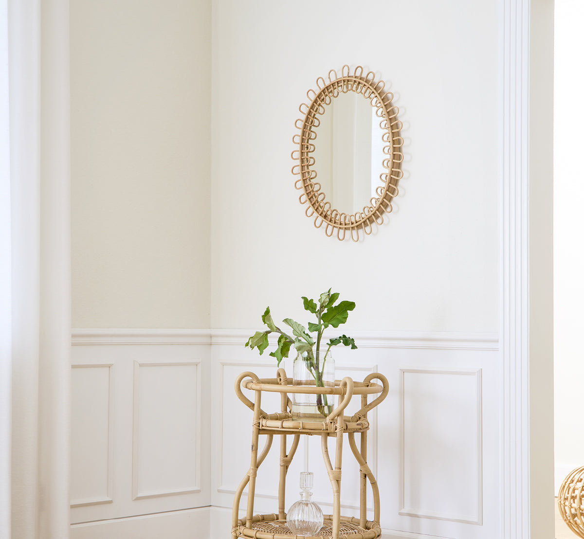 Dies ist ein Lifestyle-Bild von Luella Spiegel in polished natural rattan. An einer weiß vertäfelten Wand über einem Rattan-Barwagen mit Grün montiert.