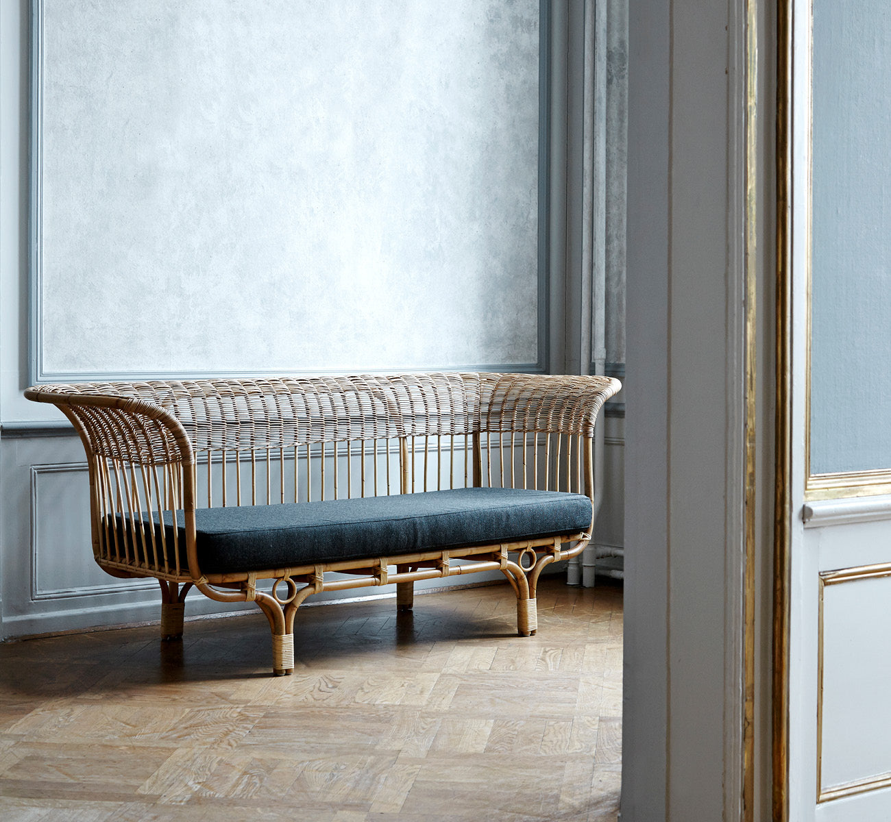 Dies ist ein Lifestyle-Bild von Belladonna Sofa in polished natural rattan, in einem hellen skandinavischen Interieur durch einen weißen Türrahmen.