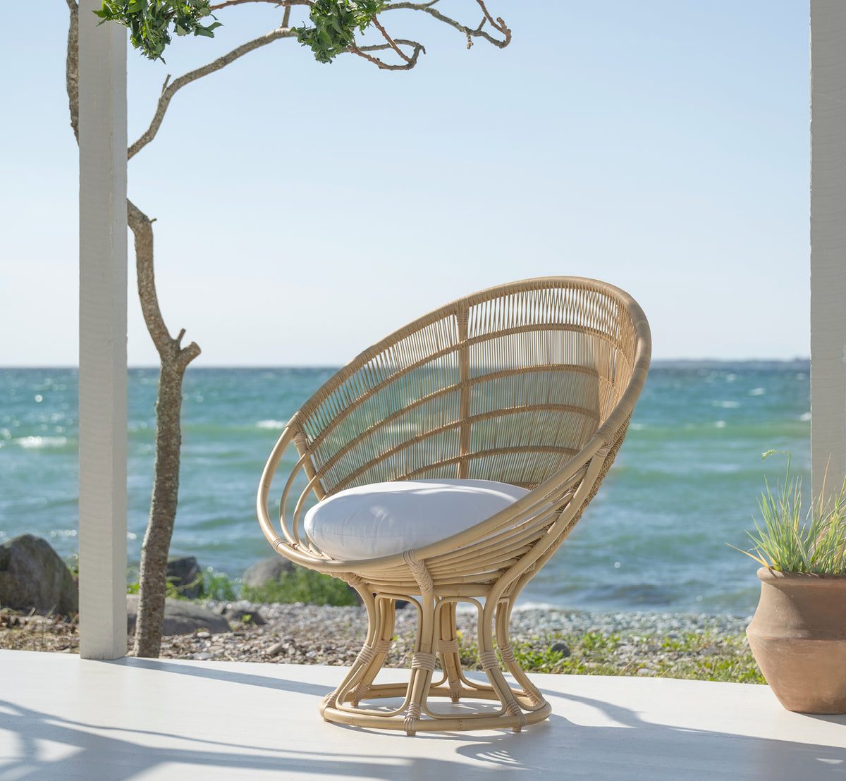 Dies ist ein Lifestyle-Bild von Luna Außen-Lounge-Sessel in natural aluminium artfibre. Auf einer weißen Strandterrasse mit Meerblick und einem Baum.