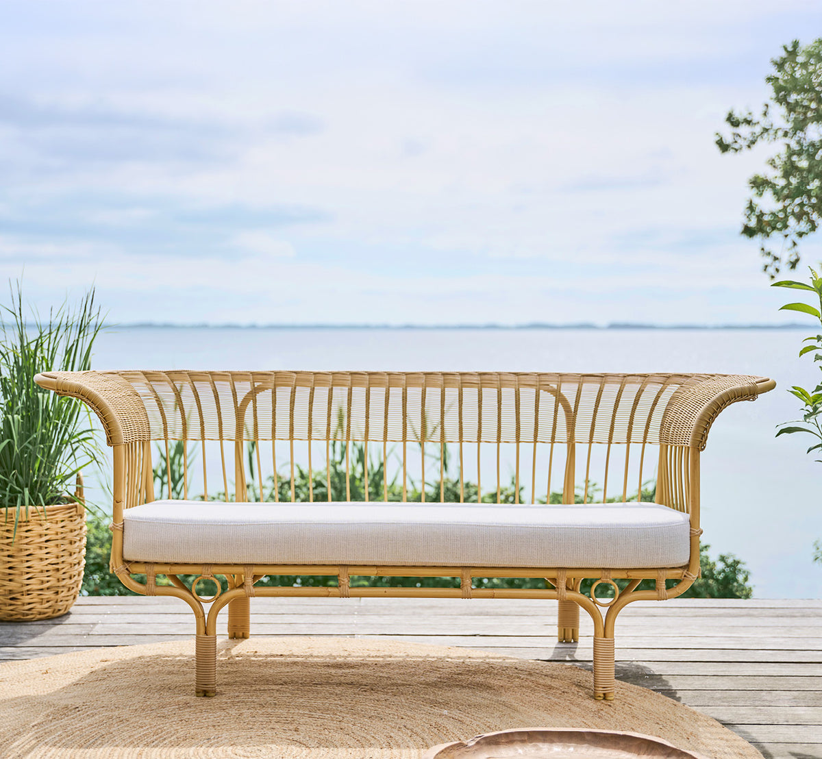 Dies ist ein Lifestyle-Bild von Belladonna Außen-Sofa in natural Aluminium ArtFibre, auf einer Holzterrasse im Freien mit Blick aufs Wasser.