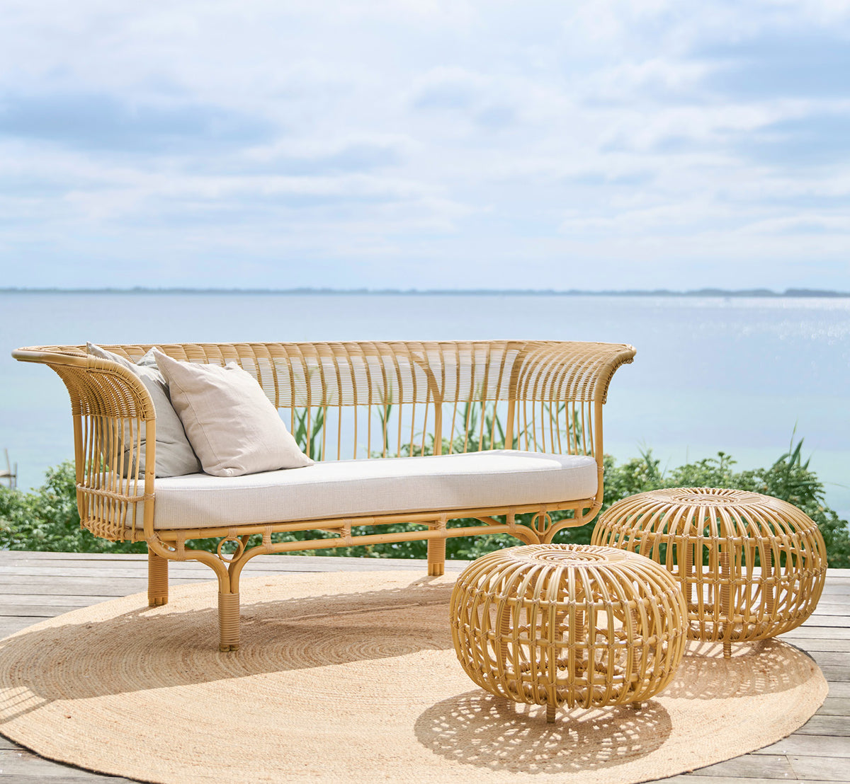 Dies ist ein Lifestyle-Bild von Belladonna Außen-Sofa in natural Aluminium ArtFibre, auf einer Holzterrasse im Freien mit Blick aufs Wasser.
