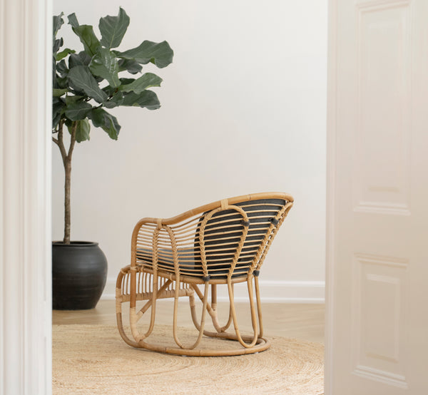 Dies ist ein Lifestyle-Bild von Swing Lounge-Sessel in polished natural rattan. Allein neben einer Grünpflanze auf einem Fischgrätparkettboden stehend.