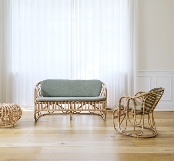 Dies ist ein Lifestyle-Bild von Swing Sofa in polished natural rattan. Auf einem Teppich neben einer Geigenfeige auf Parkett platziert.