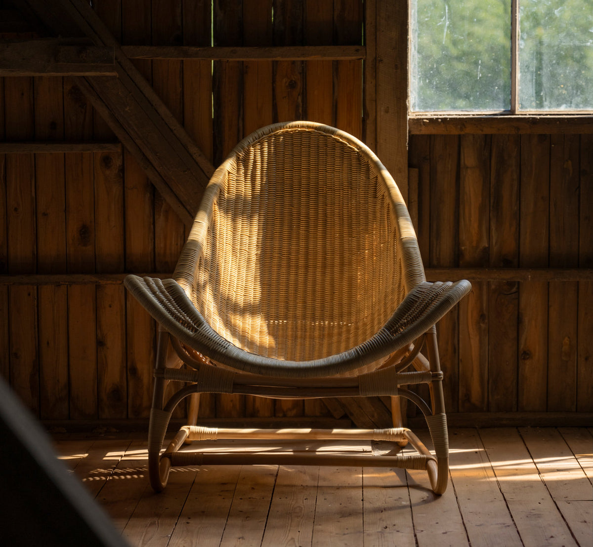 Dies ist ein Lifestyle-Bild von Pacifique Lounge-Sessel in natural core weave rattan frame. In einer Holzscheune mit warmem Fensterlicht.
