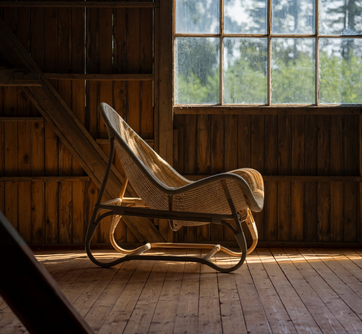 Dies ist ein Lifestyle-Bild von Pacifique Lounge-Sessel in natural core weave rattan frame. In einer Holzscheune mit warmem Fensterlicht.