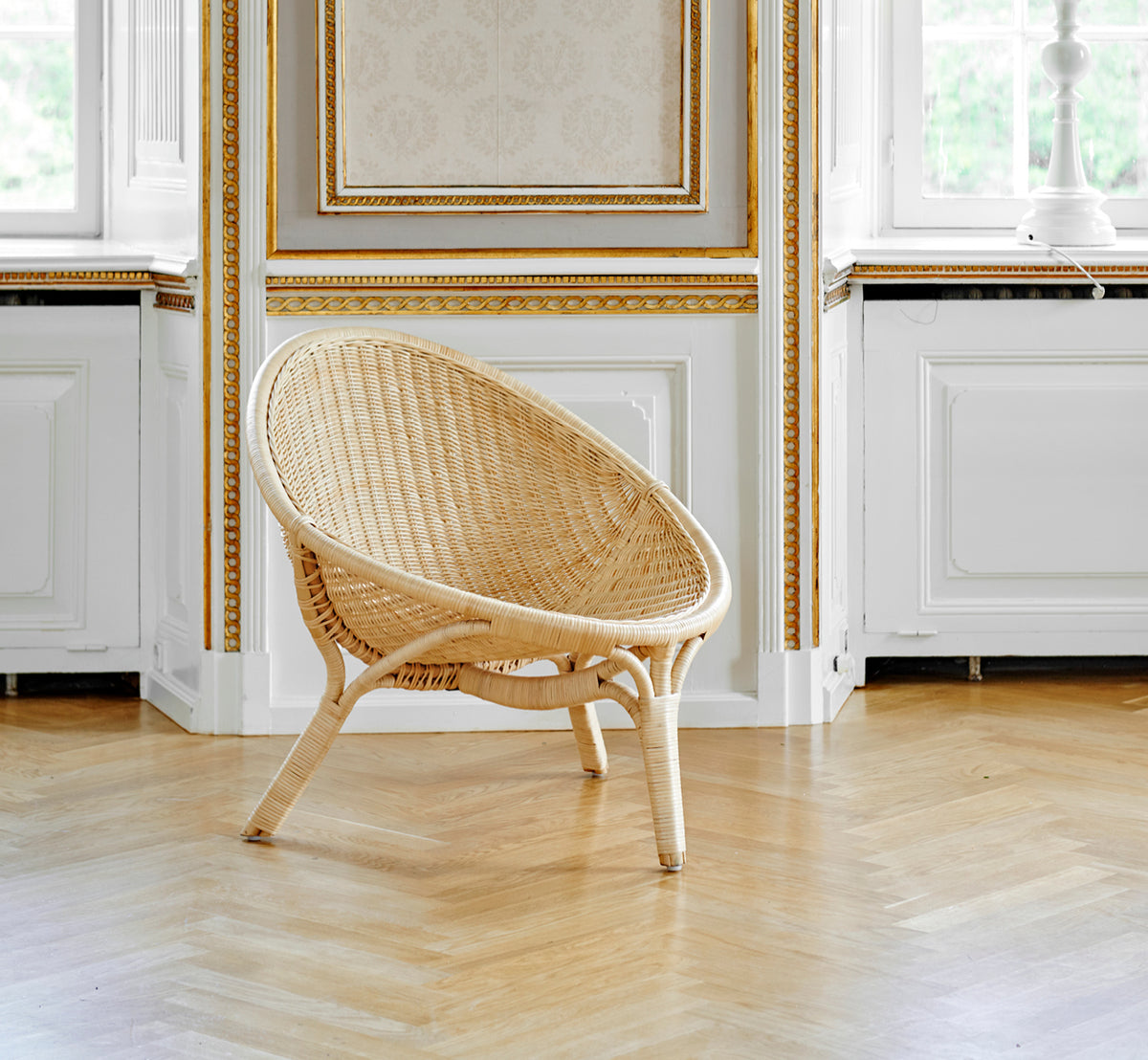 Dies ist ein Lifestyle-Bild von Rana Lounge-Sessel in natural core weave rattan frame. Allein in einem Raum mit vergoldeter Vertäfelung und Parkettboden stehend.