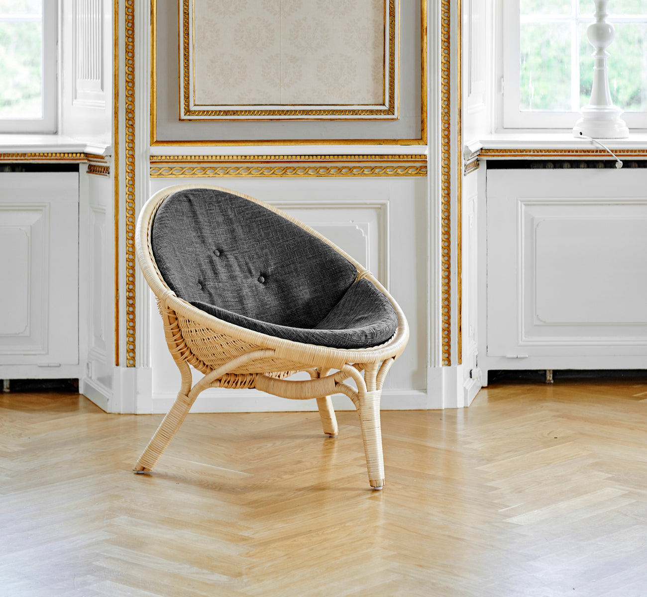 Dies ist ein Lifestyle-Bild von Rana Lounge-Sessel in natural core weave rattan frame. Allein in einem Raum mit vergoldeter Vertäfelung und Parkettboden stehend.