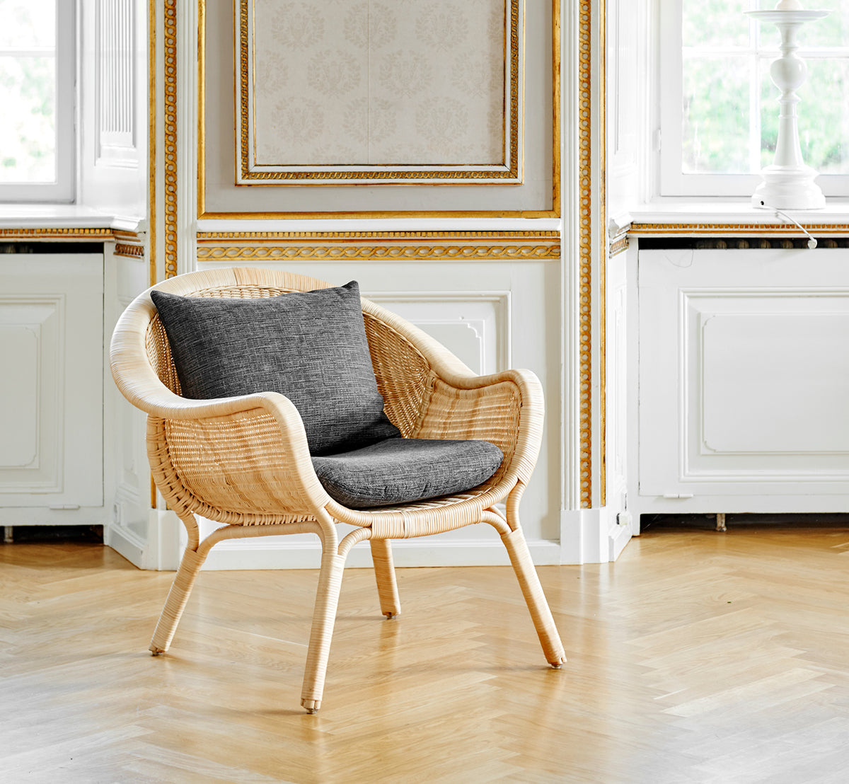 Dies ist ein Lifestyle-Bild von Madame Lounge-Sessel in natural core weave rattan frame. Eleganter vergoldeter Raum mit Eichenboden und grauem Kissen.
