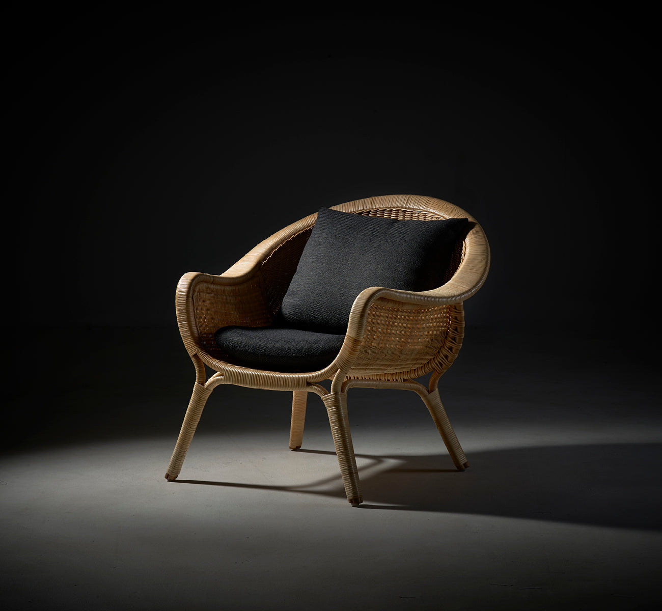 Dies ist ein Lifestyle-Bild von Madame Lounge-Sessel in natural core weave rattan frame. Dramatische Studiobeleuchtung mit dunklem Kissen vor grauem Hintergrund.