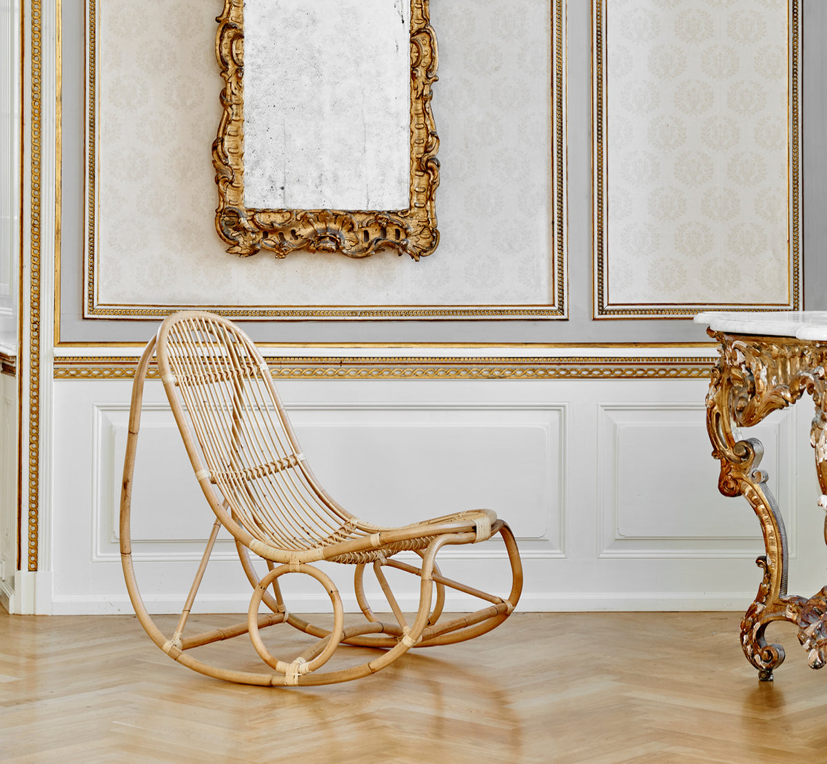 Dies ist ein Lifestyle-Bild von Nanny Rocking Chair i polished natural rattan. In einem prunkvollen vergoldeten Raum mit Barockspiegel und Konsole.