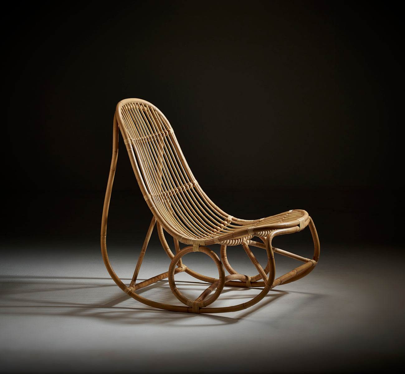 Dies ist ein Lifestyle-Bild von Nanny Rocking Chair i polished natural rattan. Studioaufnahme vor dunklem Hintergrund, die die Kurven hervorhebt.