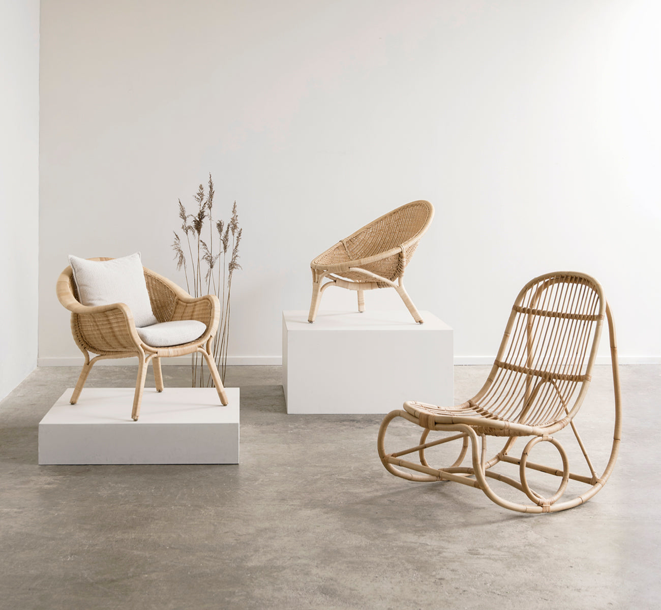 Dies ist ein Lifestyle-Bild von Nanny Rocking Chair i polished natural rattan. In einer Galerie mit Rattanstühlen auf weißen Ausstellungssockeln.