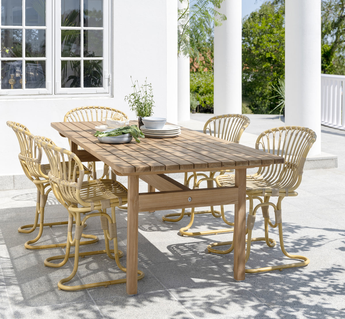 Dies ist ein Lifestyle-Bild von Rita Außen-Esszimmerstuhl in natural aluminium artfibre. Mit einem Bistrotisch auf einer weißen Kolonnadenterrasse kombiniert.