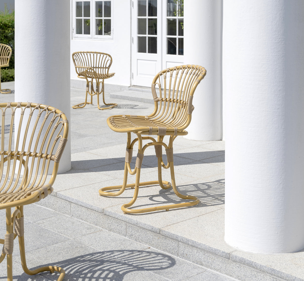 Dies ist ein Lifestyle-Bild von Rita Außen-Esszimmerstuhl in natural aluminium artfibre. Mit einem Bistrotisch auf einer weißen Kolonnadenterrasse kombiniert.