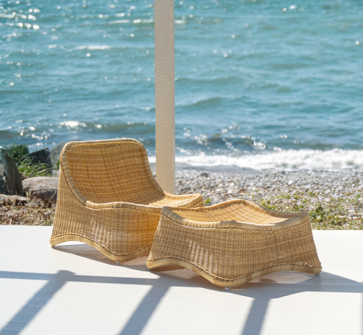 Dies ist ein Lifestyle-Bild von Chill Lounge-Sessel in natural core weave ArtFibre Aluminium, auf einem Holzdeck am Wasser mit Küstengrün und.