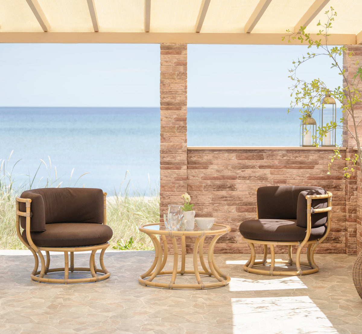 Dies ist ein Lifestyle-Bild von Coco Außen-Lounge-Sessel in natural aluminium artfibre. Lounge-Sessel und Tisch auf einer Terrasse am Meer.