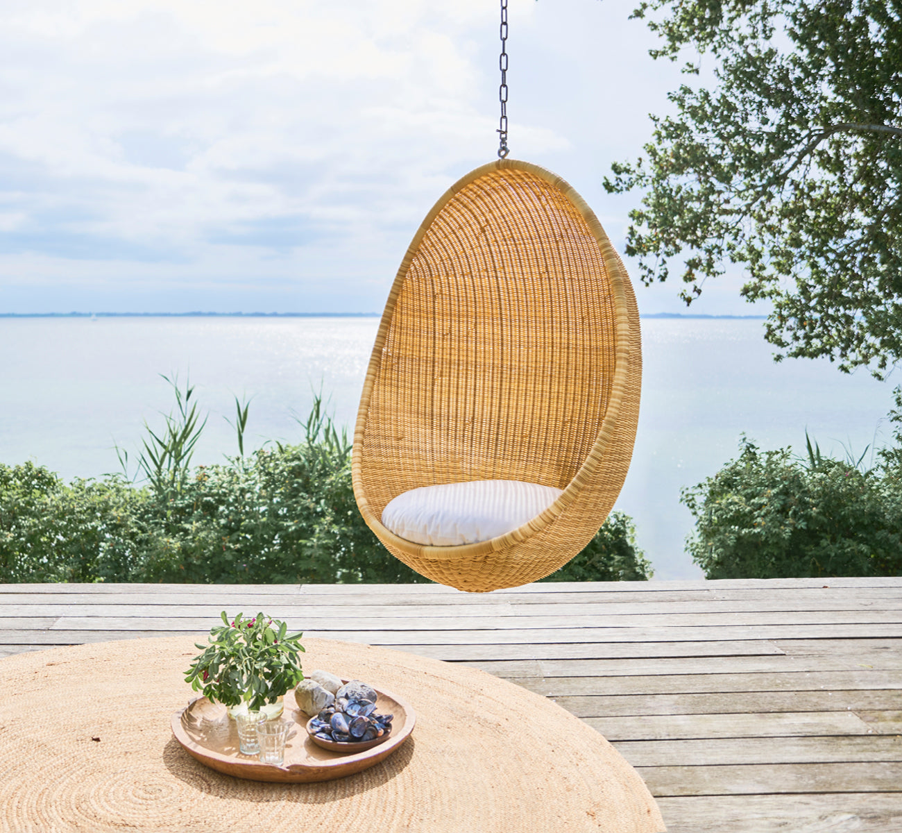 Dies ist ein Lifestyle-Bild von Hanging Außen-Hängesessel in natural core weave ArtFibre Aluminium, hängend an einer weißen Pergola mit Sandstrand.