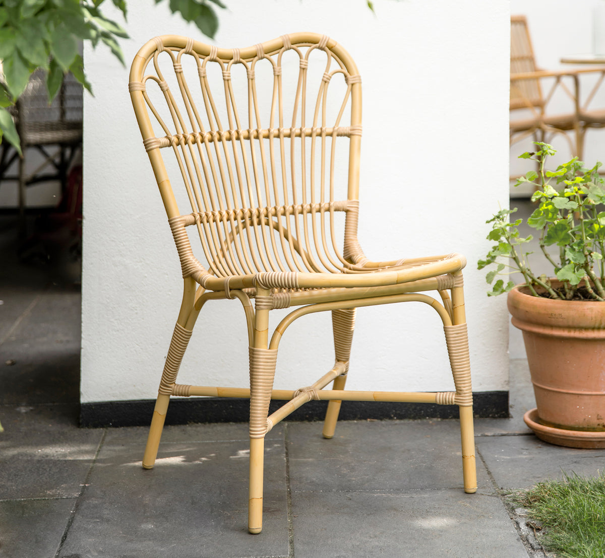 Dies ist ein Lifestyle-Bild von Margret Außen-Esszimmerstuhl in natural aluminium artfibre. Weißer Innenhof mit Terrakottatöpfen und Pflanzen.