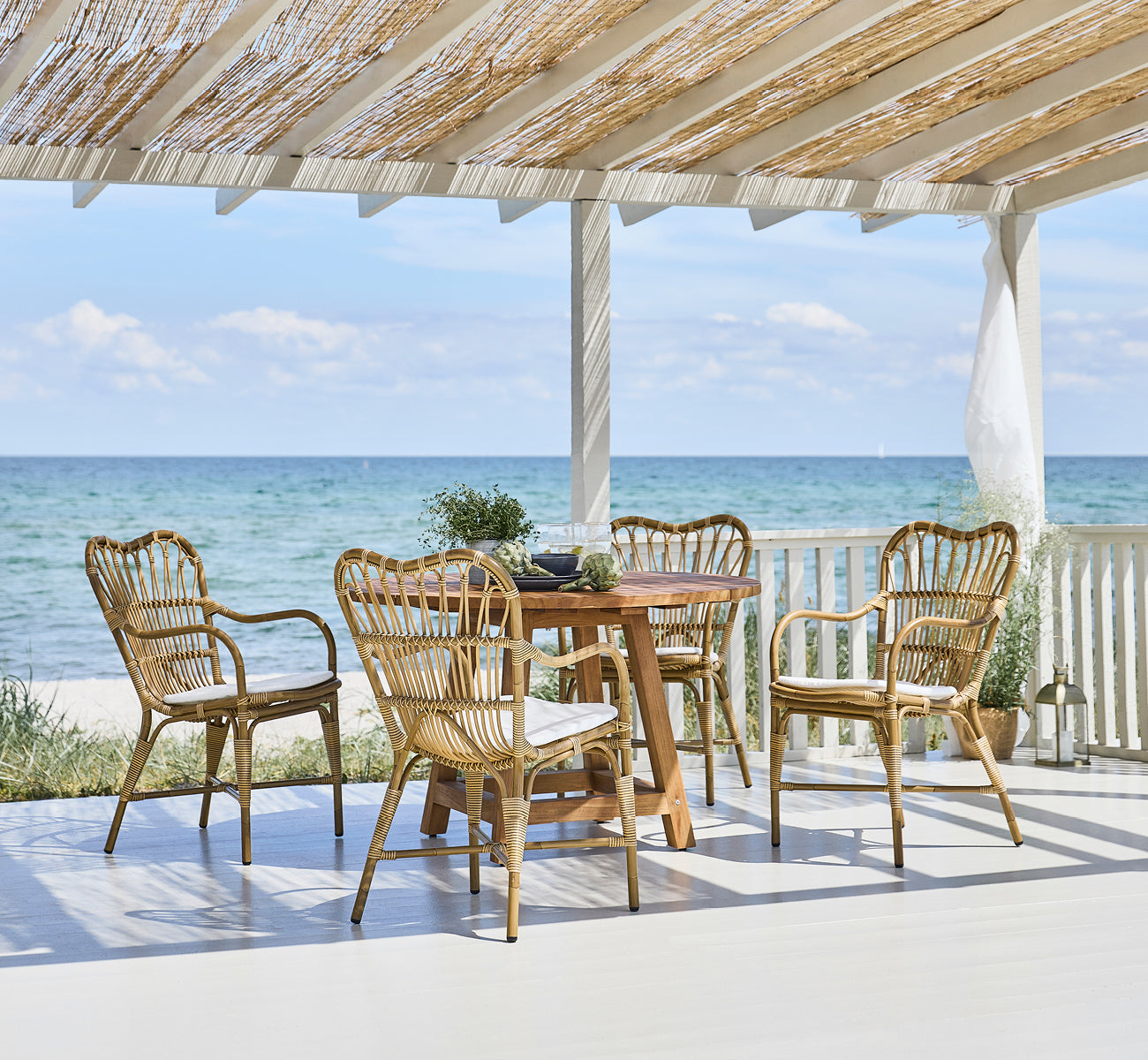 Dies ist ein Lifestyle-Bild von Margret Außen-Esszimmer-Armlehnstuhl in antique aluminium artfibre. Strandpavillon mit Teaktisch und Meerblick.