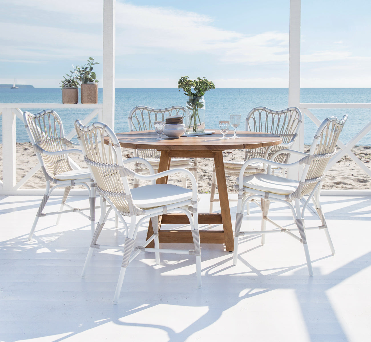 Dies ist ein Lifestyle-Bild von Margret Außen-Esszimmer-Armlehnstuhl in dove white aluminium artfibre. Strandterrasse mit Teaktisch und Meerblick.