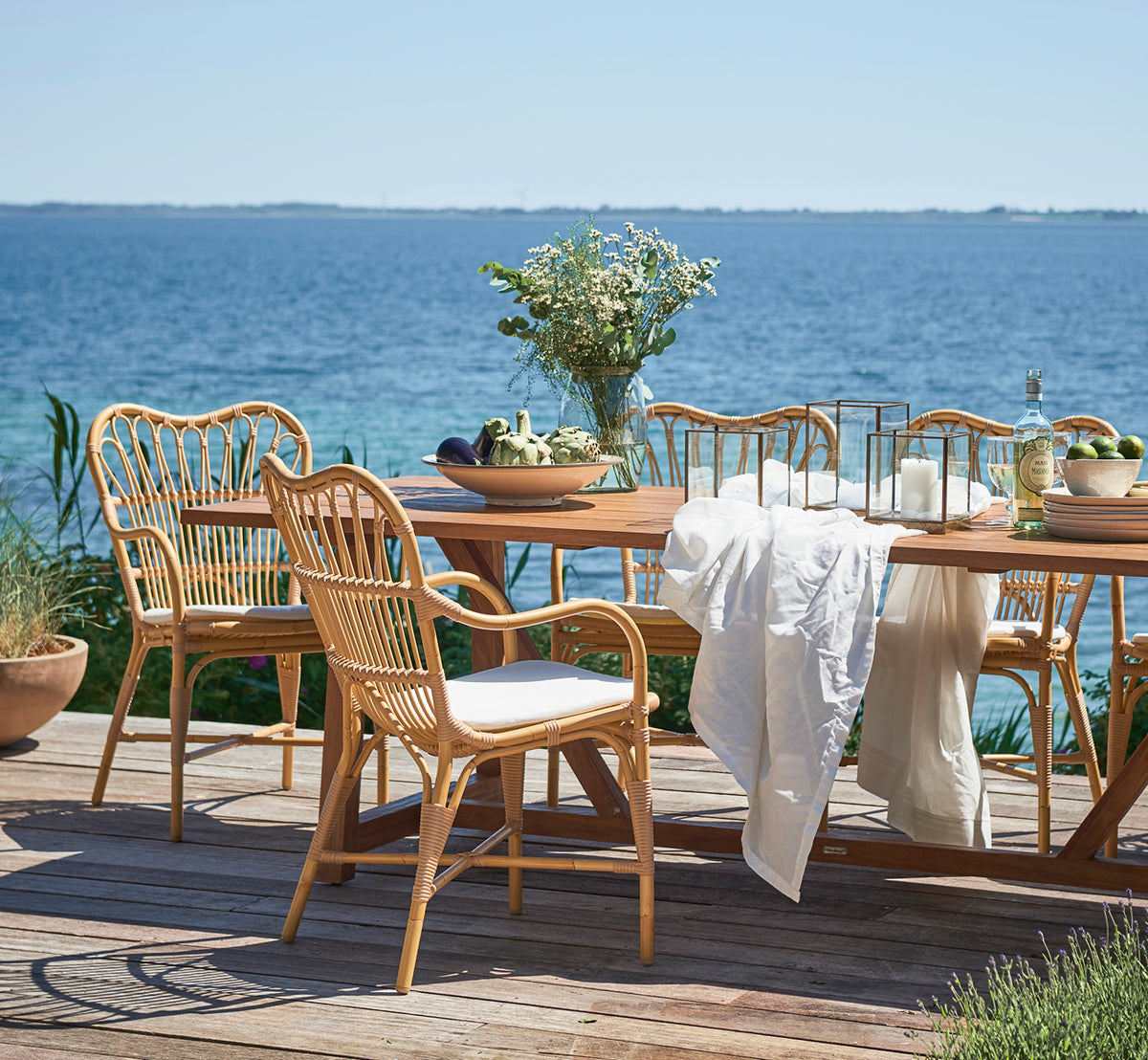 Dies ist ein Lifestyle-Bild von Margret Außen-Esszimmer-Armlehnstuhl in natural aluminium artfibre. Holzterrasse am Meer mit Teaktisch und Blumen.