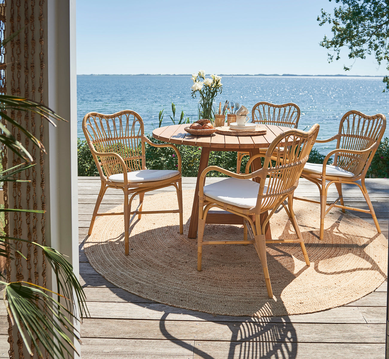 Dies ist ein Lifestyle-Bild von Margret Außen-Esszimmer-Armlehnstuhl in natural aluminium artfibre. Terrasse am Meer mit rundem Teaktisch und Meerblick.