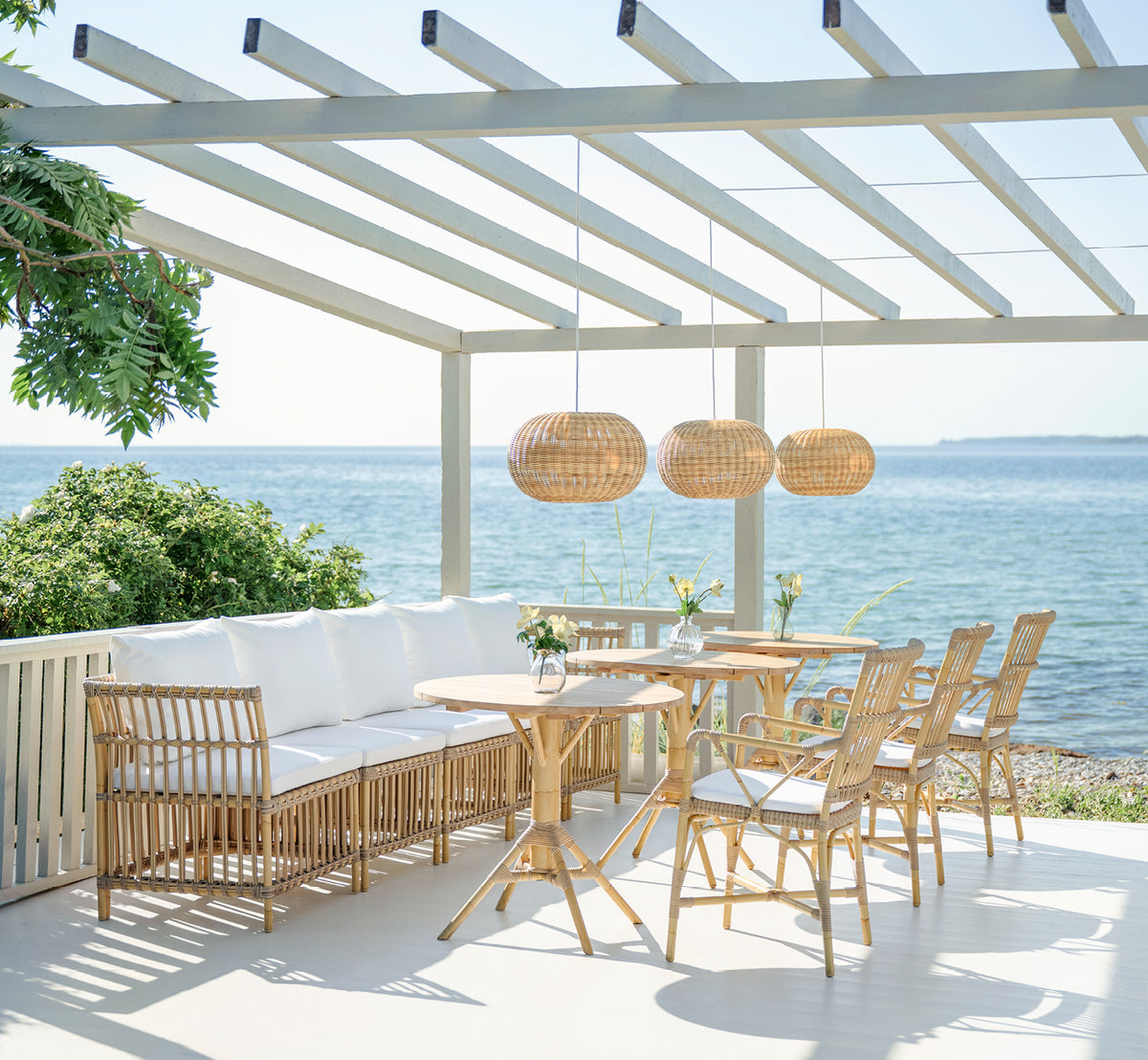 Dies ist ein Lifestyle-Bild von Piano Exterior Dining Armchair i natural aluminium artfibre. Unter einer Pergola am Meer mit Rattan-Pendelleuchten.