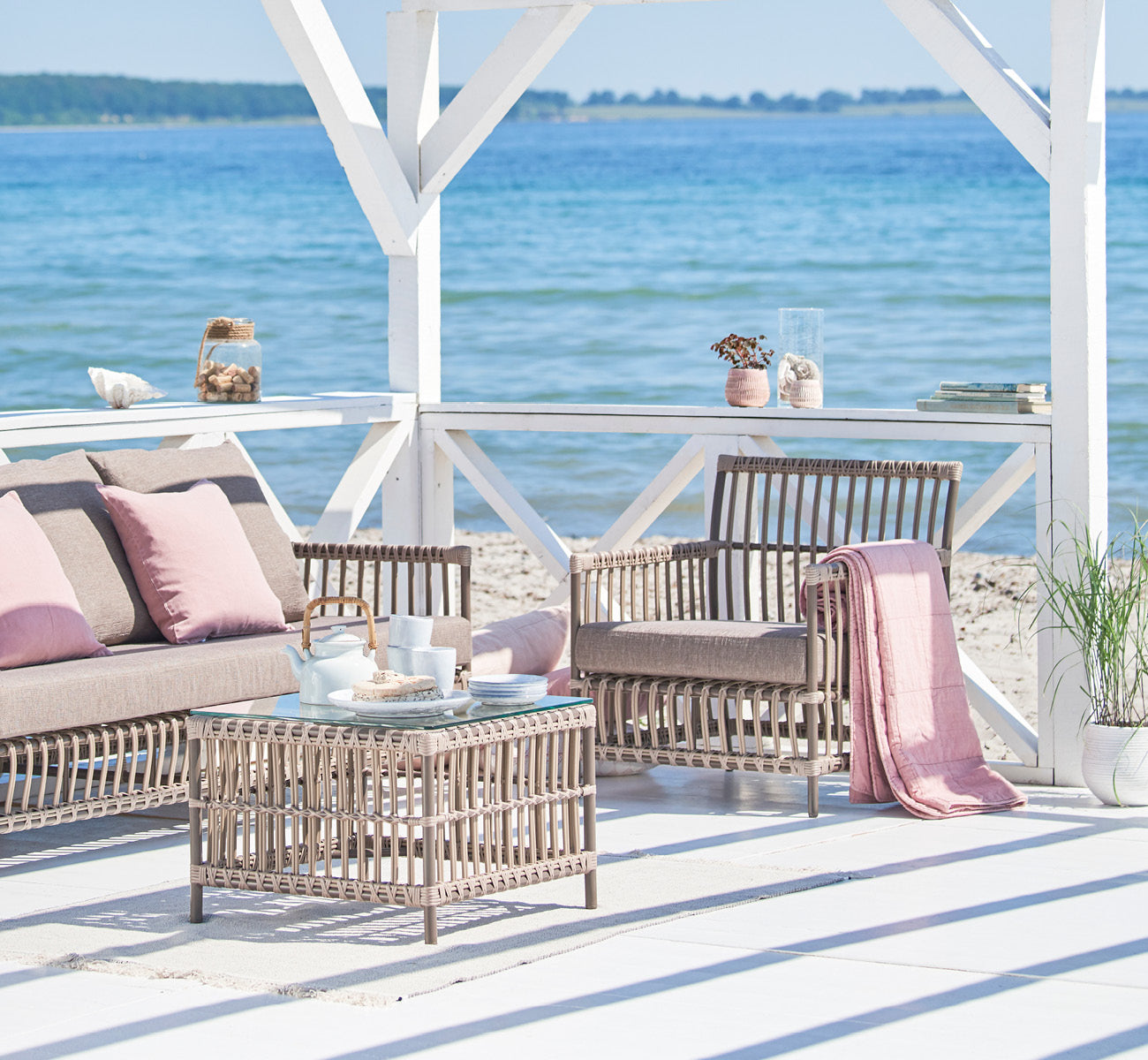 Dies ist ein Lifestyle-Bild von Caroline Außen-Lounge-Sessel in moccaccino Aluminium ArtFibre, auf einem Holzdeck am Wasser mit Küstengrün und.