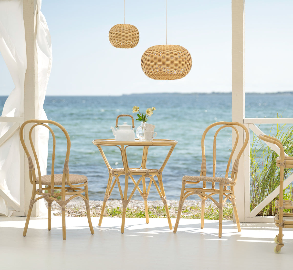 Dies ist ein Lifestyle-Bild von Lulu Außen-Esszimmerstuhl in natural aluminium artfibre. Auf einer Strandterrasse mit einem runden Tisch und Meerblick.