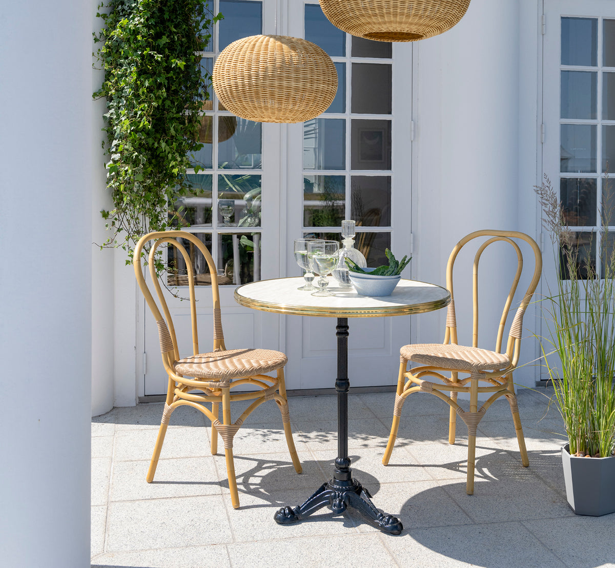 Dies ist ein Lifestyle-Bild von Lulu Außen-Esszimmerstuhl in natural aluminium artfibre. Auf einer weißen Terrasse mit einem Cafétisch und Kletterpflanzen.