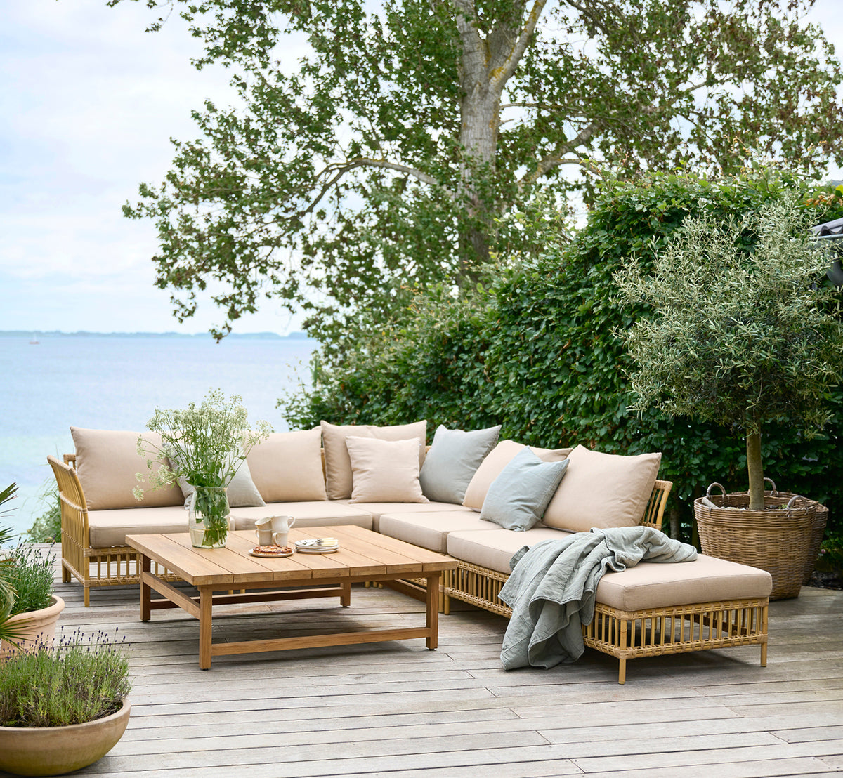 Dies ist ein Lifestyle-Bild von Maggie Sofa Eckmodul in natural aluminium artfibre. Holzterrasse am Meer mit Kissen und Meerblick.