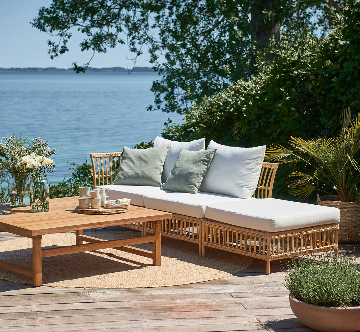 Dies ist ein Lifestyle-Bild von Maggie Sofa Eckmodul in natural aluminium artfibre. Terrasse am Meer mit Couchtisch und grünen Kissen.