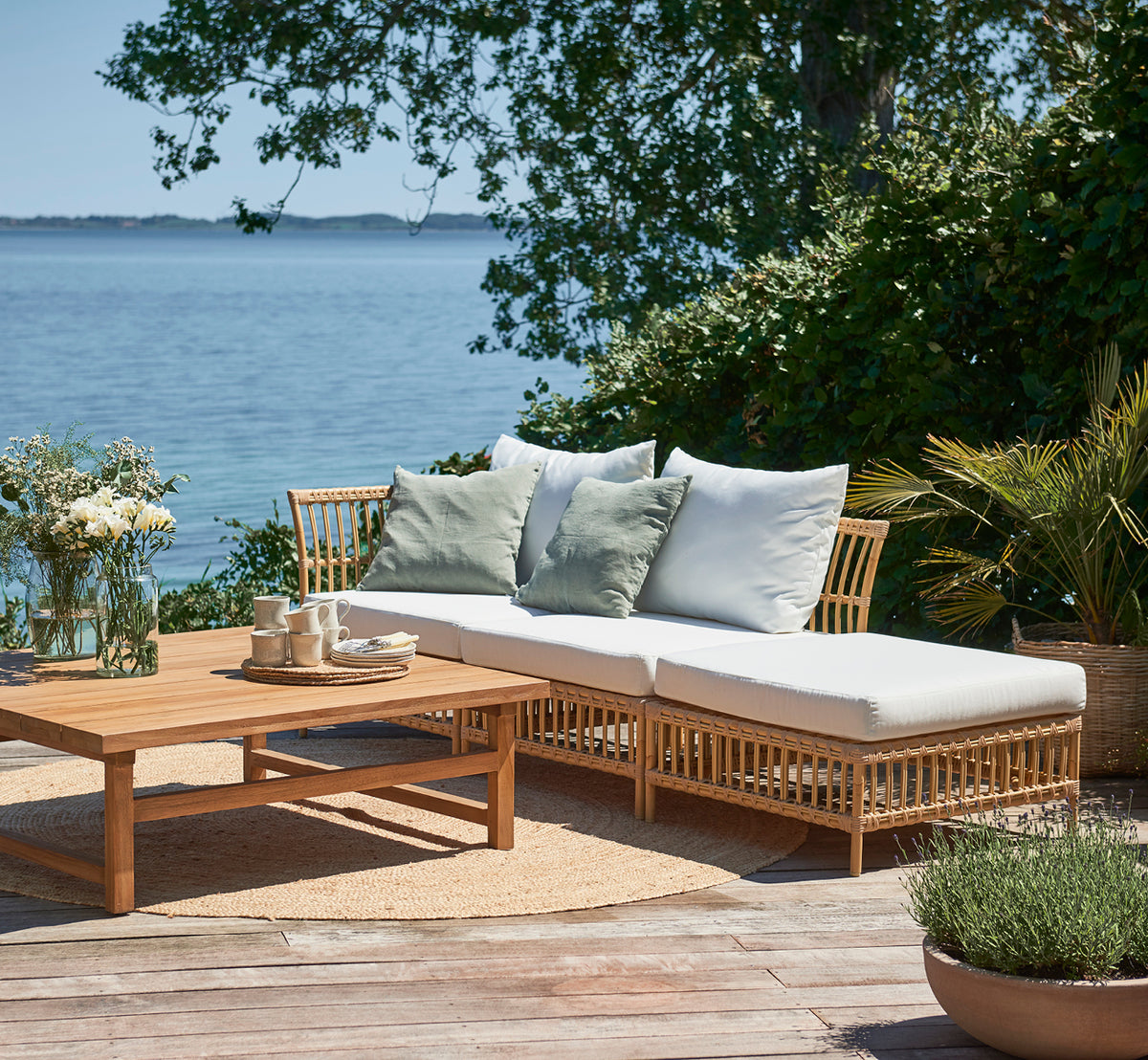 Dies ist ein Lifestyle-Bild von Maggie Sofa Mittelmodul in natural aluminium artfibre. Terrasse am Meer mit Kissen, Pflanzen und Meerblick.