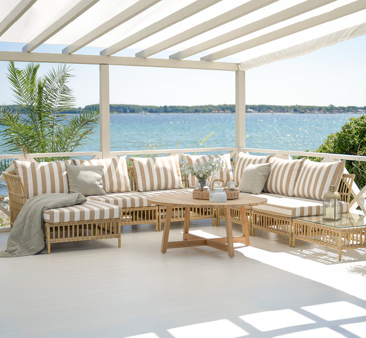Dies ist ein Lifestyle-Bild von Maggie Sofa Mittelmodul in natural aluminium artfibre. Überdachte Terrasse mit gestreiften Kissen und Meerkulisse.