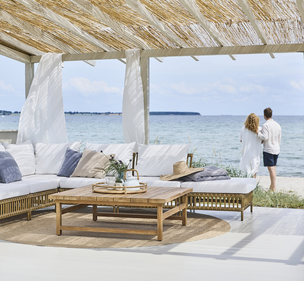 Dies ist ein Lifestyle-Bild von Maggie Sofa Pouf-Modul m. Glas in antique aluminium artfibre. Strandpavillon mit Paar und Meerblick.