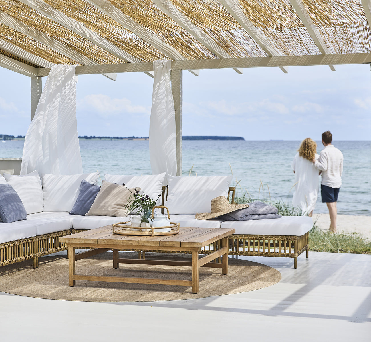 Dies ist ein Lifestyle-Bild von Maggie Sofa Pouf-Modul m. Glas in antique aluminium artfibre. Strandpavillon mit Paar und Meerblick.