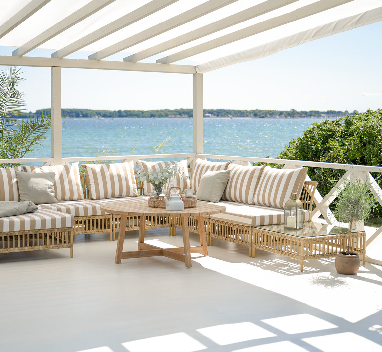 Dies ist ein Lifestyle-Bild von Maggie Sofa Pouf-Modul m. Glas in natural aluminium artfibre. Überdachte Terrasse mit gestreiften Kissen und Meerblick.