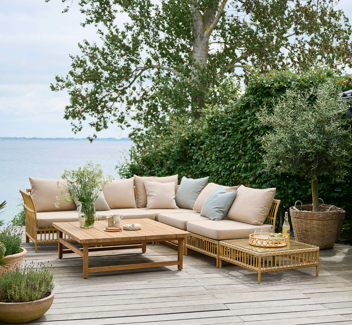Dies ist ein Lifestyle-Bild von Maggie Sofa Pouf-Modul m. Glas in natural aluminium artfibre. Terrasse am Meer mit Sofaset und Grün drumherum.