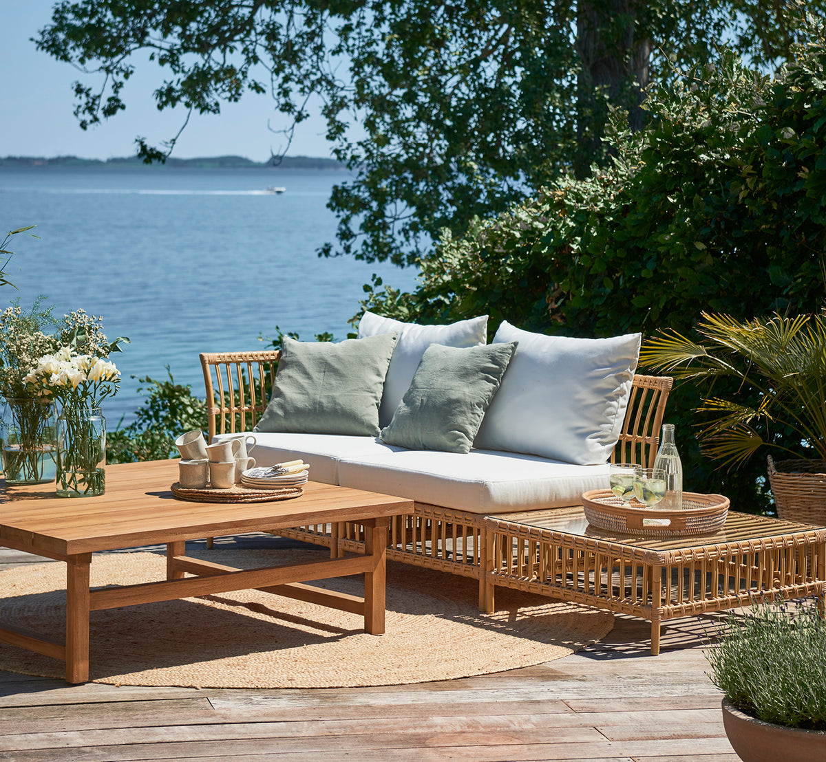 Dies ist ein Lifestyle-Bild von Maggie Sofa Pouf-Modul m. Glas in natural aluminium artfibre. Terrasse am Meer mit Couchtisch und grünen Kissen.