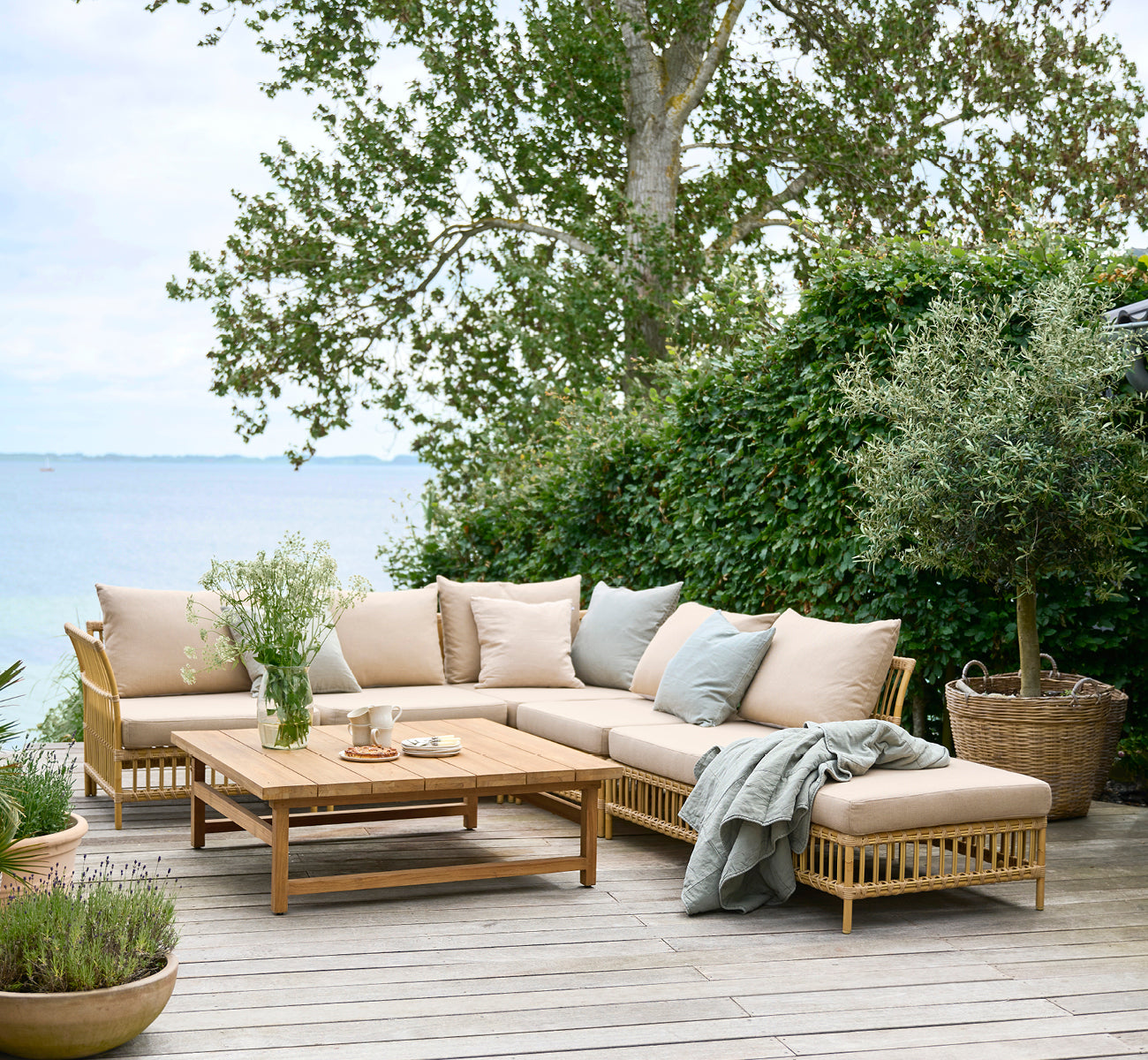 Dies ist ein Lifestyle-Bild von Maggie Sofa Pouf-Modul m. Glas in natural aluminium artfibre. Terrasse am Meer mit Modulsofa und drapierter Decke.