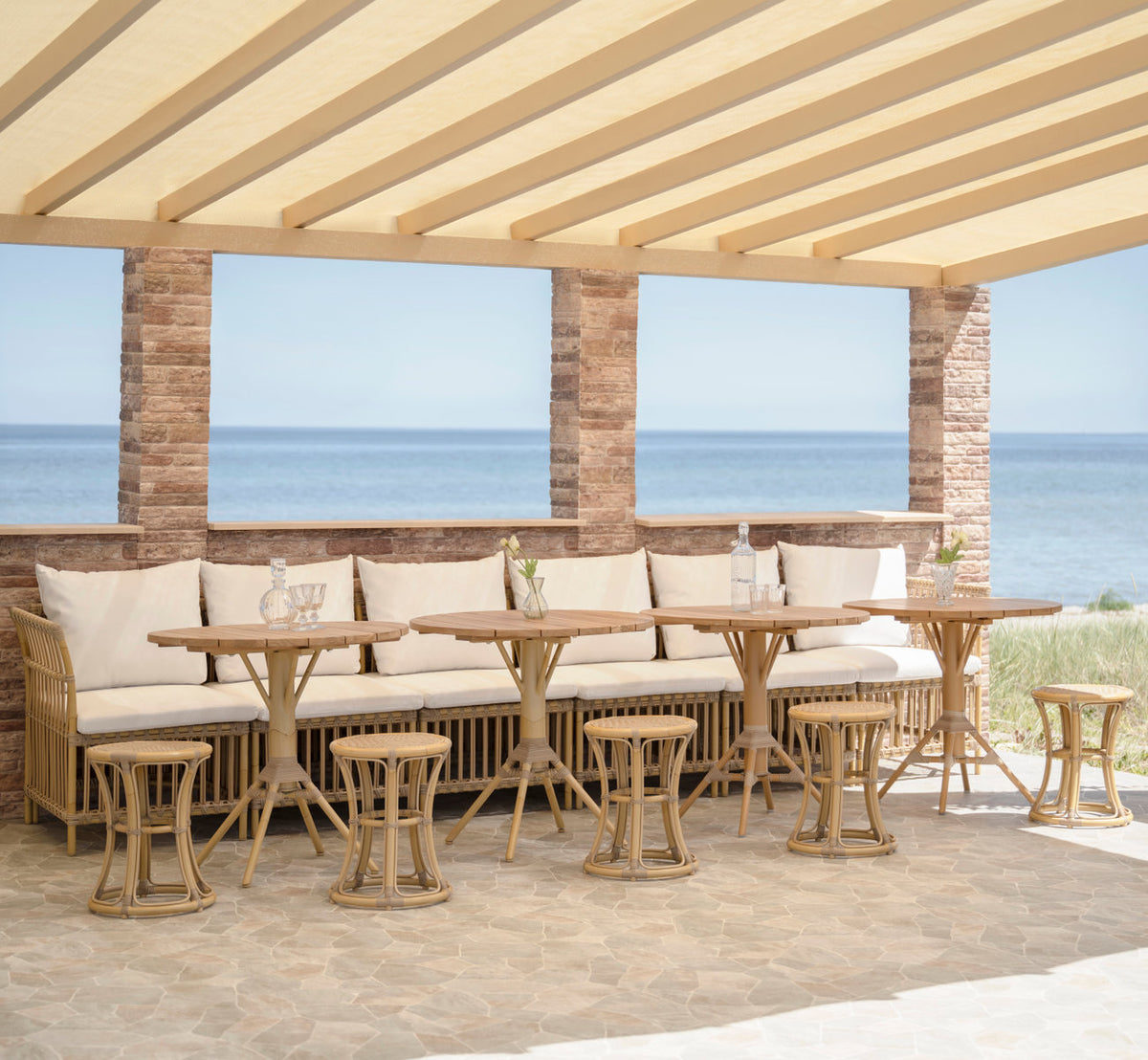 Dies ist ein Lifestyle-Bild von Meghan Außen-Essbank-Corner Module in natural aluminium artfibre. Unter einer Pergola am Meer.