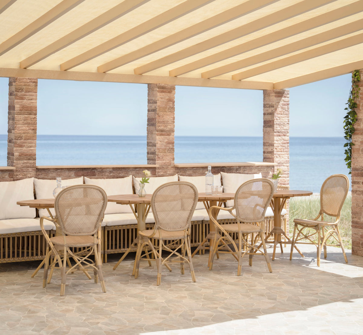 Dies ist ein Lifestyle-Bild von Meghan Außen-Essbank-Center Module in natural aluminium artfibre. Unter einer Pergola am Meer.