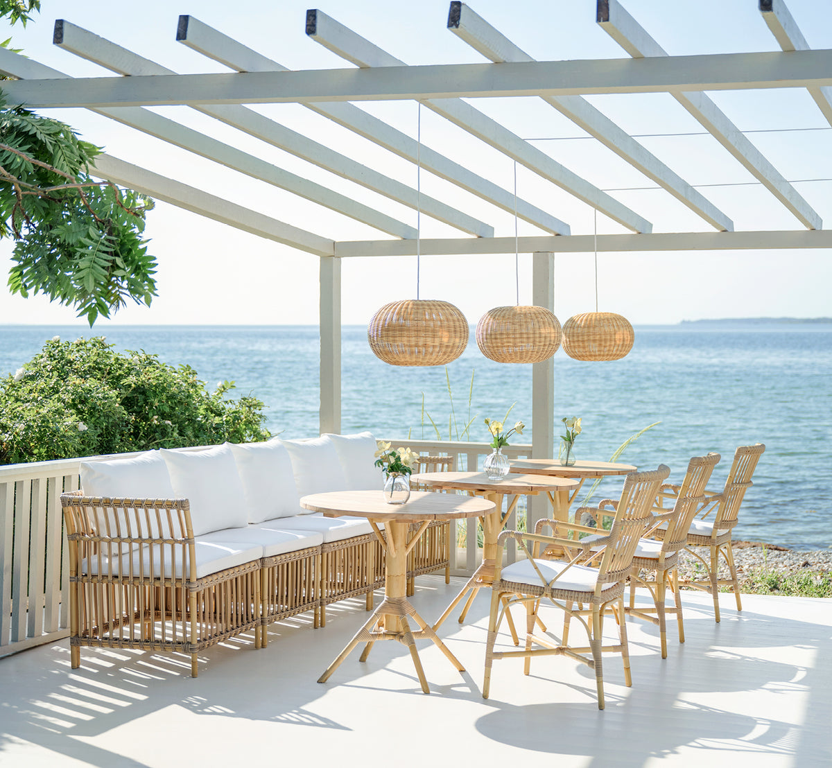 Dies ist ein Lifestyle-Bild von Meghan Exterior Dining Bench Center Module i natural aluminium artfibre. Auf einem Pergola-Deck am Meer.