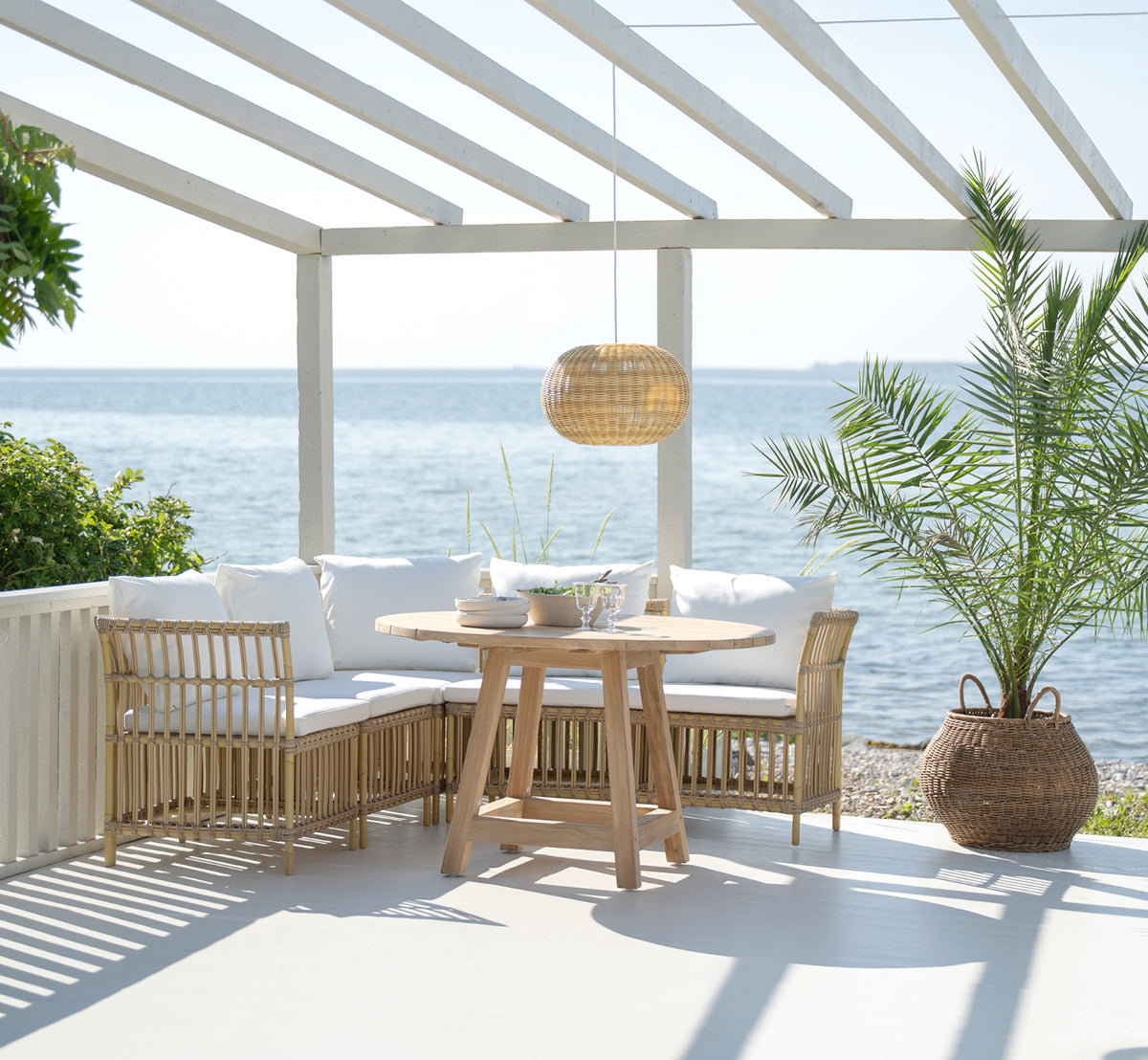 Dies ist ein Lifestyle-Bild von Meghan Exterior Dining Bench Center Module i natural aluminium artfibre. Um einen runden Tisch am Meer.
