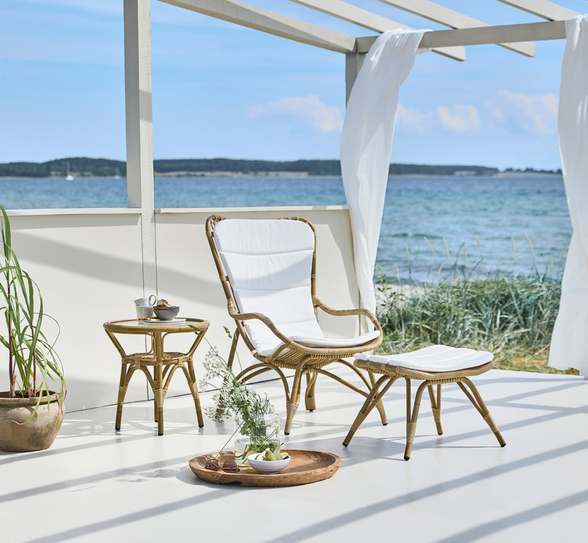 Dies ist ein Lifestyle-Bild von Monet Exterior Lounge Chair i antique aluminium artfibre. Mit Fußhocker auf einem Deck in einem Pavillon am Meer.