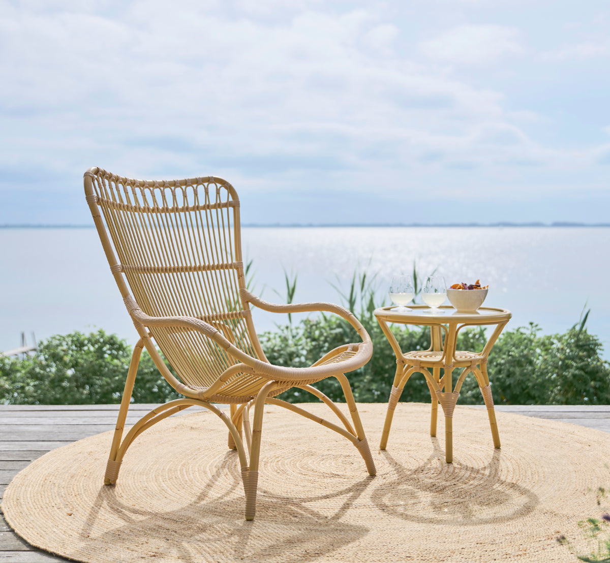 Dies ist ein Lifestyle-Bild von Monet Exterior Lounge Chair i natural aluminium artfibre. Auf einem Holzdeck mit Juteteppich und Meerblick.