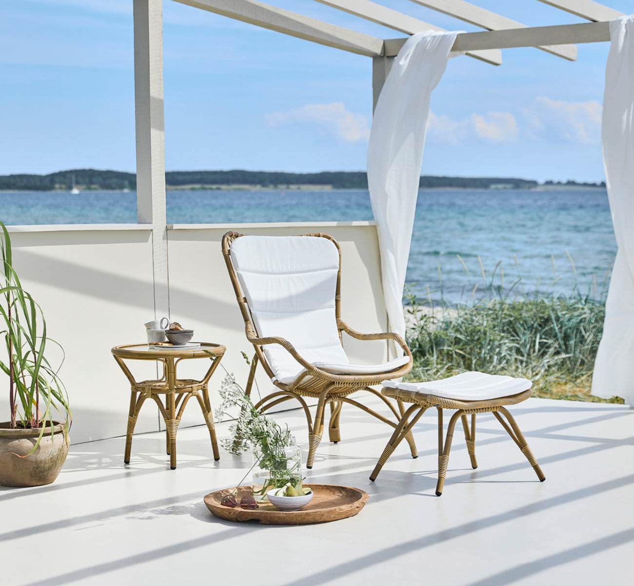 Dies ist ein Lifestyle-Bild von Monet Exterior Footstool i antique aluminium artfibre. In einem Pavillon am Meer mit gepolstertem Sessel.