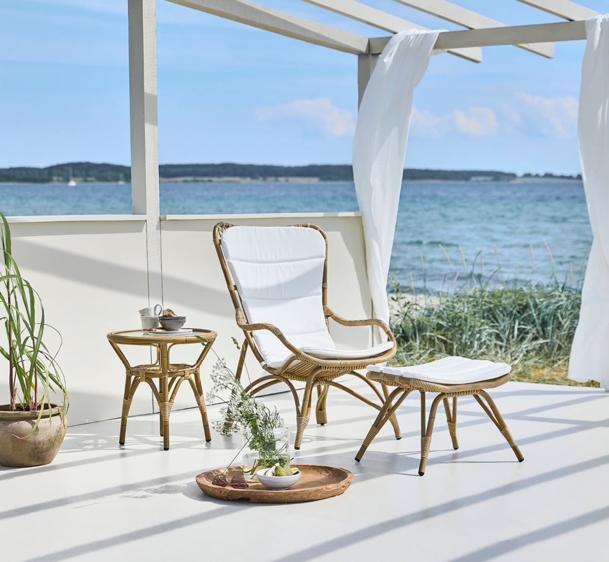 Dies ist ein Lifestyle-Bild von Monet Exterior Footstool i antique aluminium artfibre. In einem Pavillon am Meer mit gepolstertem Sessel.