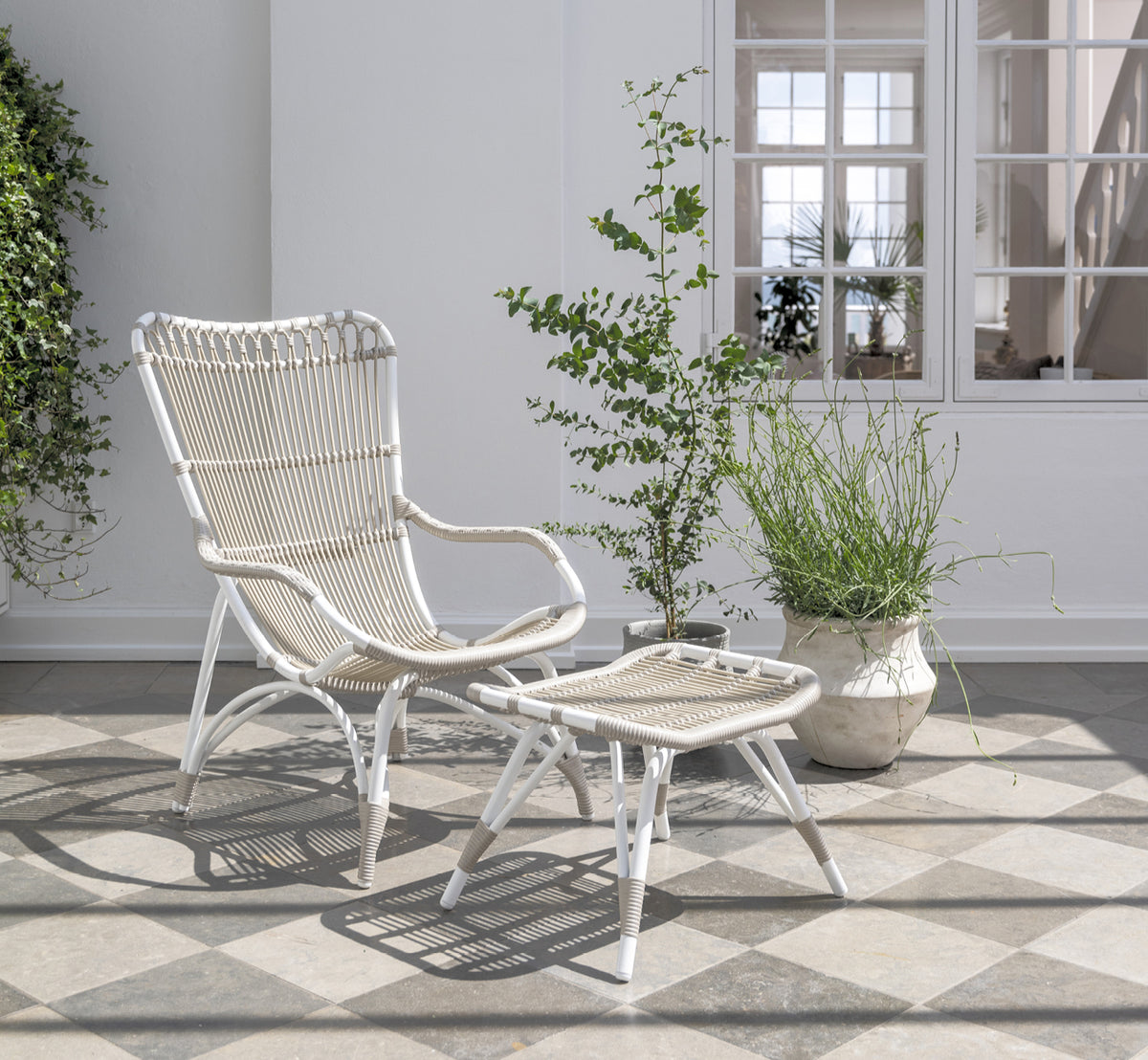 Dies ist ein Lifestyle-Bild von Monet Exterior Footstool i dove white aluminium artfibre. Auf einer Strandveranda mit Lounge-Sessel.